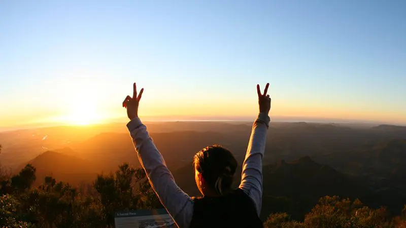 Person triumphantly raising arms at sunrise atop mountain after Hinterland Magic Glow Worm Experience, celebrating scenic adventure.