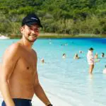 Happy man in swimming trunks enjoys a scenic beach on his 27-day tour, with crystal-clear blue water and swimmers in the background.