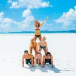 A group of six people builds a human pyramid on a sunny, sandy East Coast beach beneath vivid blue, cloud-speckled skies.
