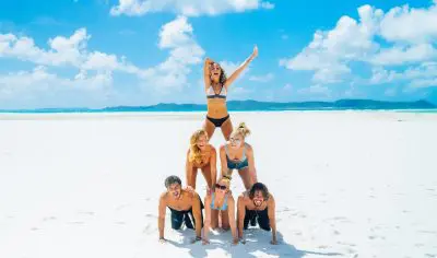 A group of six people builds a human pyramid on a sunny, sandy East Coast beach beneath vivid blue, cloud-speckled skies.