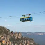 Blue and yellow cable car glides over dramatic Blue Mountains cliffs on a top-rated 1 Day Tour under clear skies, showcasing iconic views.