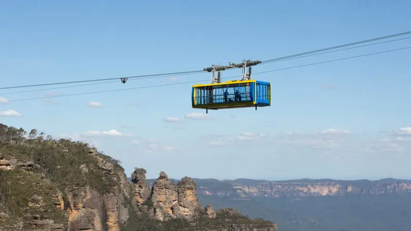 Blue and yellow cable car glides over dramatic Blue Mountains cliffs on a top-rated 1 Day Tour under clear skies, showcasing iconic views.