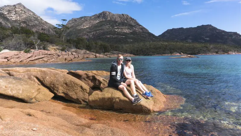 Tourists relax on a scenic rock overlooking crystal-clear blue waters during the Wineglass Bay Full Day Tour via Richmond, Tasmania.