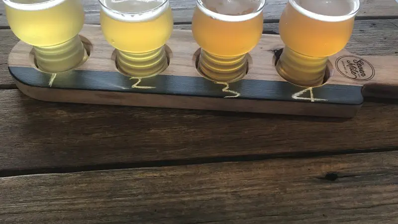 Craft beer tasting flight with four glasses in a wooden holder on a rustic table, ideal for Arvo Session Behind The Barrel tours.