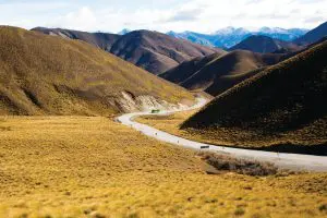 Image of A scenic winding road that snakes through golden-brown grassy hills with majestic mountains beyond, beneath a vibrant partly cloudy sky as part of Kiwi Experience South Island Adventure