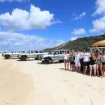 Adventure seekers stand next to rugged off-road vehicles on a sandy beach beneath a vibrant blue sky with fluffy white clouds.