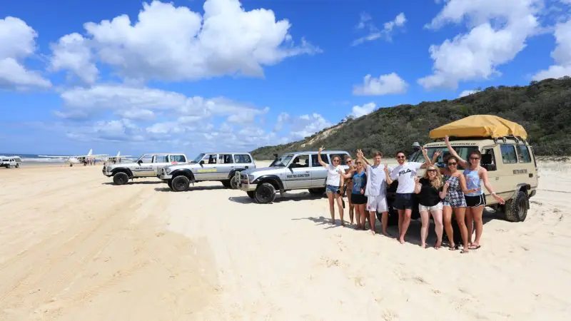 Happy group posing on Fraser Island’s sandy beach next to 4WD vehicles during a top-rated 2 Day Kgari Tag Along Adventure with Dingoes.