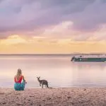 Woman enjoying a sunset on Noosa Everglades beach beside a kangaroo after a 1 Day Explorer tour, sandy shore in the background.