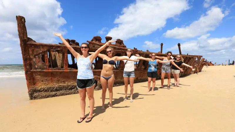 Group of six women on sandy Fraser Island beach during 2 Day K'gari adventure tour, shipwreck and blue sea in the background.