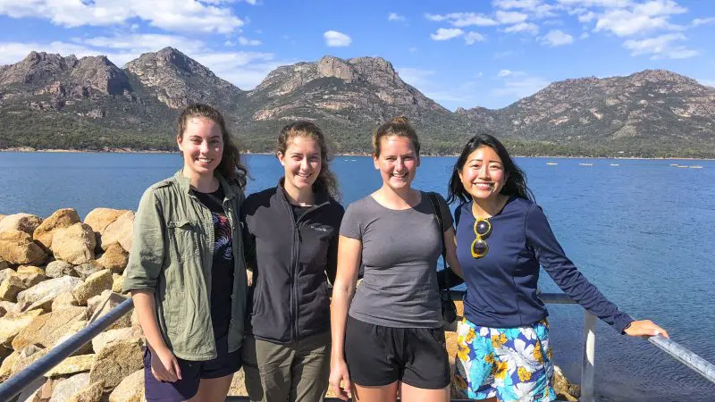 Four women smile by the crystal-blue lake at Wineglass Bay on a full-day tour via Richmond, scenic mountains and clouds in the background.