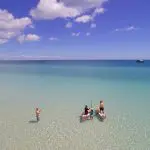 Tourists paddleboarding in crystal-clear, shallow waters during the Remote 1 Day K'gari Fraser Island tour under a blue, clouded sky.
