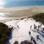 Tour group experiences Remote 1 Day K'gari Fraser Island adventure on pristine white sand dunes beside blue ocean under sunny skies.