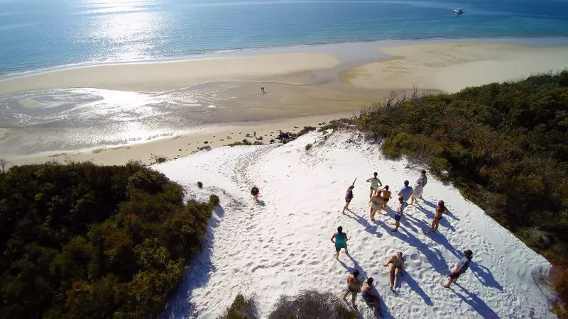 Tour group experiences Remote 1 Day K'gari Fraser Island adventure on pristine white sand dunes beside blue ocean under sunny skies.