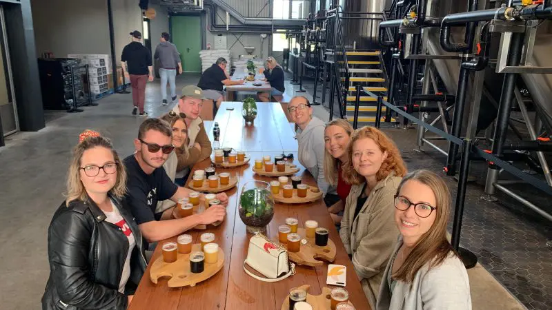 Enthusiastic group samples beer flights at Arvo Session Behind The Barrel Half Day Tour, smiling together at a communal brewery table.