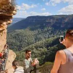 Tourists take photos on a Blue Mountains Day Tour with scenic waterfall walk and group lunch, overlooking lush forested valley views.