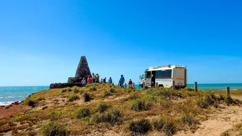 Tourists on a Kakadu Arnhem Land Tour gather by a stone monument and coach atop a grassy hill with panoramic sea views.