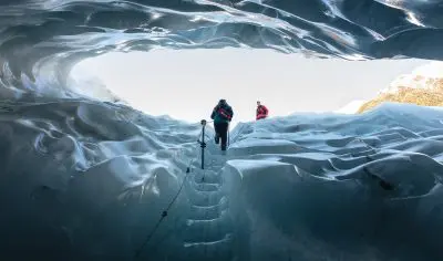 Two adventurers ascend an icy staircase inside Franz Josef Glacier, enveloped by shimmering blue ice walls and glowing natural skylight.