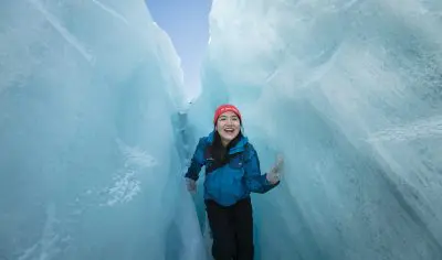 A smiling adventurer in winter gear stands surrounded by striking blue ice walls on a Franz Josef Glacier guided tour in New Zealand.