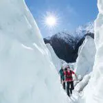 Three hikers trek Franz Josef Glacier, navigating towering ice walls under clear skies, encircled by dramatic snowy mountain peaks.