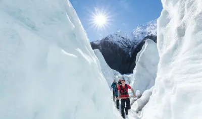 Three hikers trek Franz Josef Glacier, navigating towering ice walls under clear skies, encircled by dramatic snowy mountain peaks.