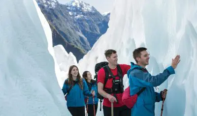 Four adventurers hike icy walls on a Franz Josef Glacier tour, smiling with joy against breathtaking New Zealand mountain scenery.