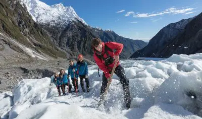 Four adventurers heli-hiking Franz Josef Glacier, one breaking pristine glacier ice with an ice axe beneath breathtaking alpine views.