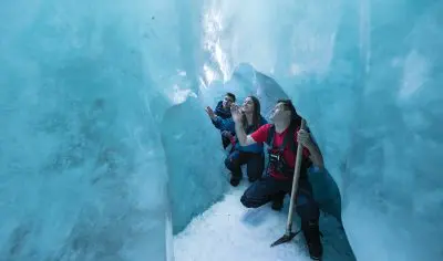 Three adventurers trek through a vivid blue Franz Josef Glacier ice cave, gazing upward in wonder as one wields an ice axe.