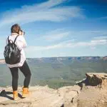 Adventurer with rucksack photographing panoramic Blue Mountains vista from rugged cliff edge on Uncharted Tour, sweeping valley below.