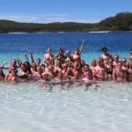 A large group in swimming costumes smiles while swimming in a lake during their 3 Day K’gari Adventure with dormitory accommodation at Rainbow Beach.
