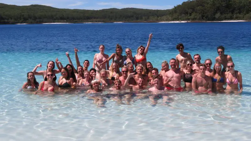 A large group in swimming costumes smiles while swimming in a lake during their 3 Day K’gari Adventure with dormitory accommodation at Rainbow Beach.