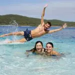 Two women enjoy crystal-clear water while a man leaps in, splashing, on a guided 3 Day K'gari Adventure tour from Rainbow Beach.
