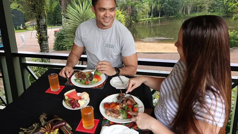 Two friends enjoy al fresco dining by a tranquil pond, smiling after a scenic journey on the Grand Kuranda Railway in Cairns.