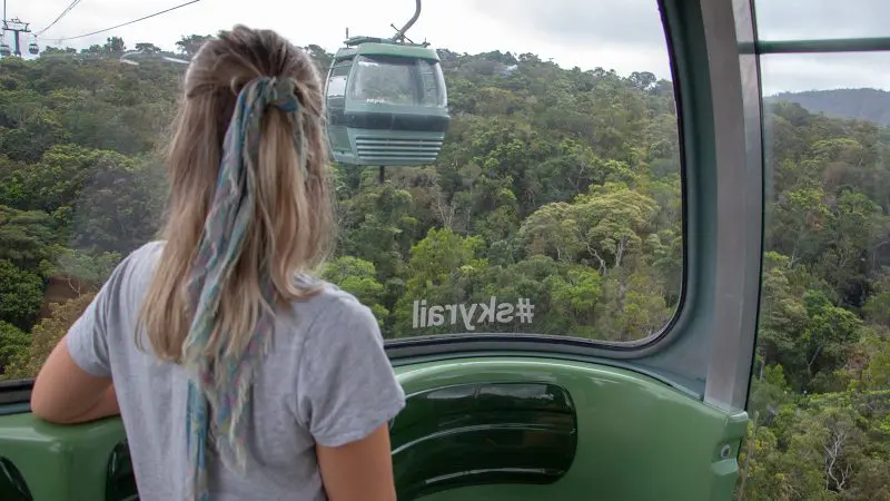 Woman with ponytail rides Skyrail cable car above vibrant rainforest, views Kuranda Scenic Railway train on famous Star Tour below.