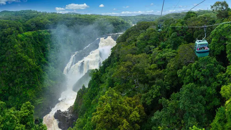 Skyrail cable car travels above vibrant rainforest canopy, overlooking a majestic waterfall under a bright blue sky with fluffy clouds.