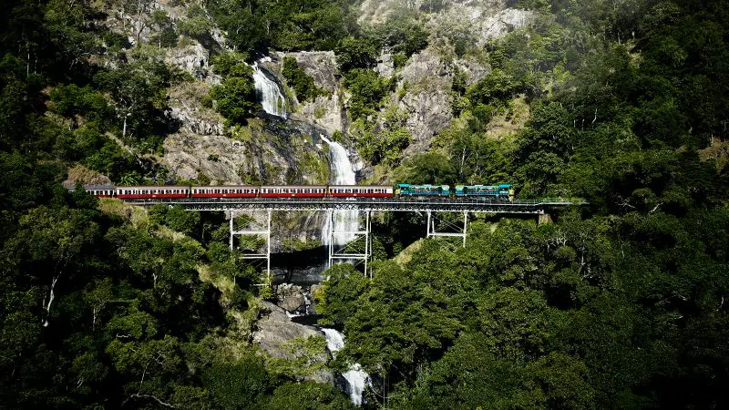 The iconic red and green Grand Kuranda train crosses a scenic bridge near a cascading waterfall, surrounded by lush rainforest.
