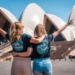Two joyful women in Ultimate Travel shirts raise their arms, celebrating the guided 7-week East Coast adventure from Melbourne to Cairns.