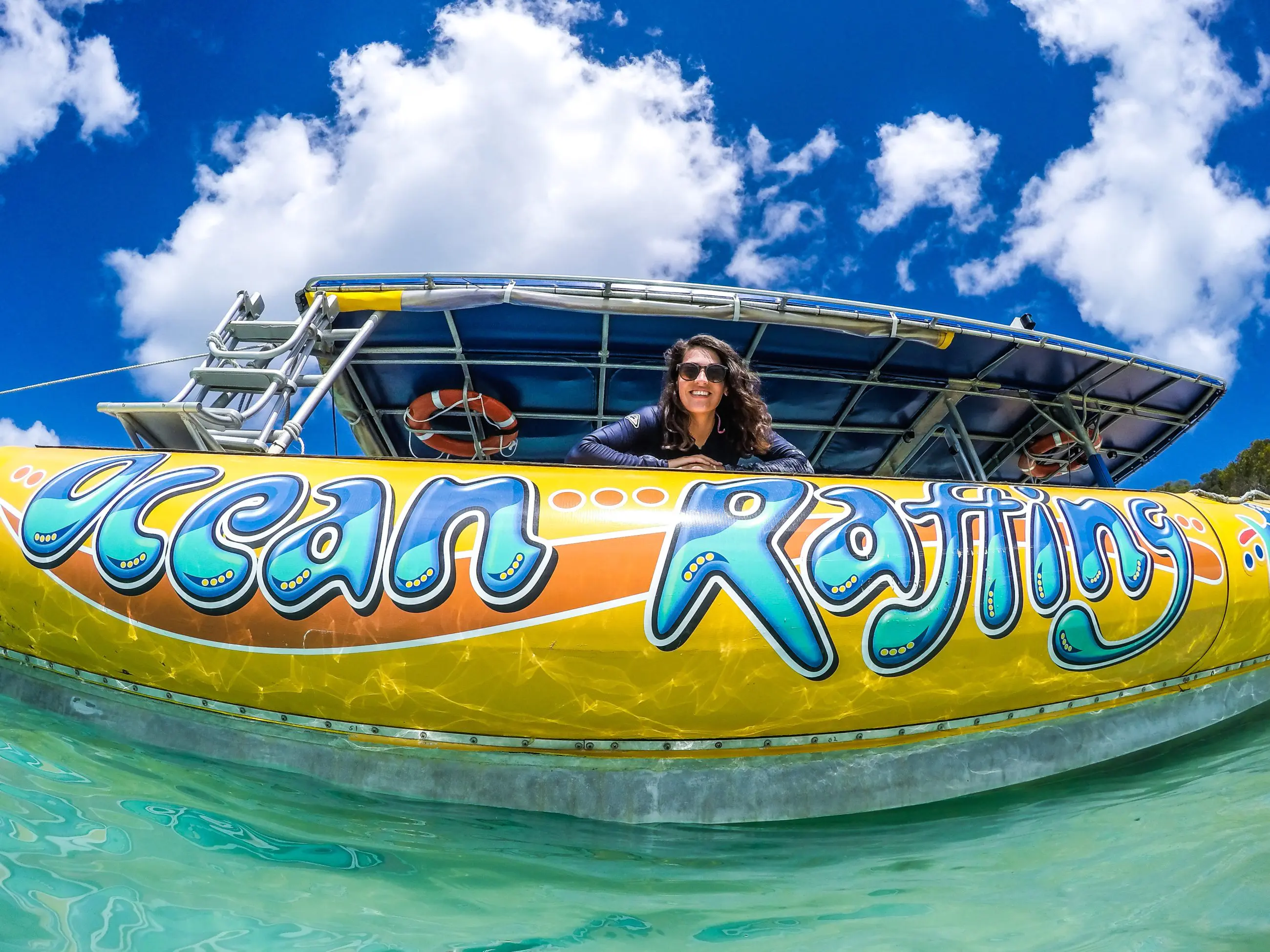 Happy woman leans over vibrant Ocean Rafting Northern Exposure Tour boat in crystal-clear water beneath a bright, blue sky with clouds.
