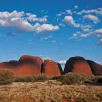 Majestic red rock formations tower above sunlit dry grass and vivid blue skies on the Uluru Rock tour from Alice Springs to Yulara.