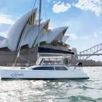 A sleek white catamaran cruises Sydney Harbour with the iconic Opera House and vibrant Vivid Lights, perfect for a BYO experience.