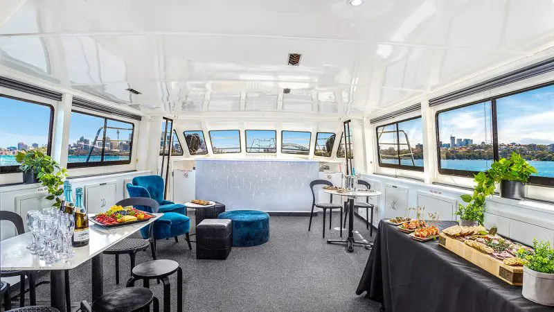 Luxurious Sydney Harbour Vivid Lights Cruise interior with gourmet canapés, plush blue seating, and breath-taking city vistas through panoramic windows.