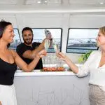 Two women toast champagne on an intimate Sydney Harbour Vivid Lights Cruise as a barman pours drinks with the illuminated city skyline.