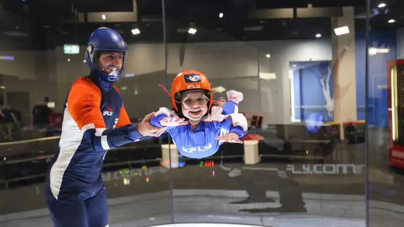 Smiling child in full flight kit experiences indoor skydiving with an instructor at iFLY Family and Friends wind tunnel attraction.