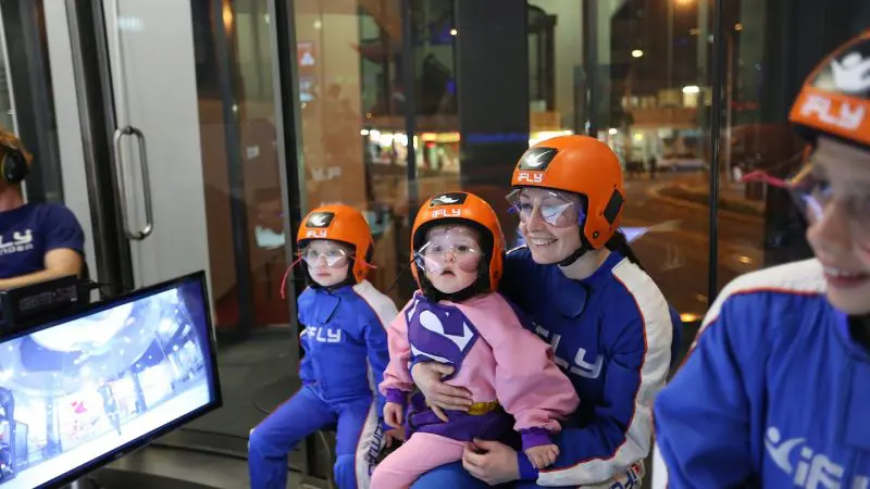 Four people in flight suits and orange helmets smile indoors at an iFLY Family and Friends skydiving session, capturing joyful moments.