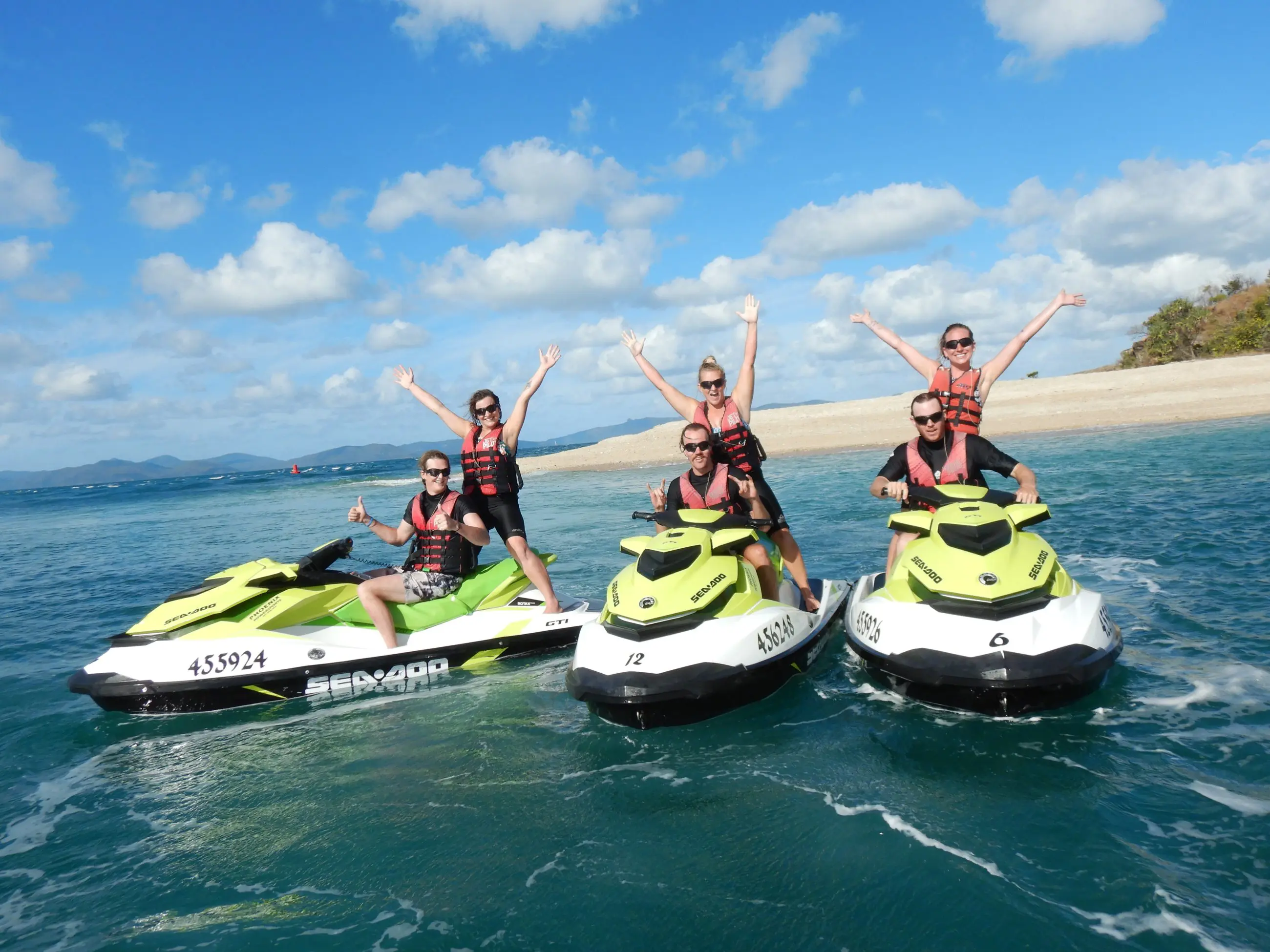 Group of five on two vibrant yellow jet skis, smiling and raising arms during Airlie Adventure Jetski Tour by a scenic sandy beach.
