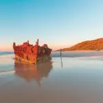 Discover the iconic rusty shipwreck on Fraser Island’s sandy beach at sunset, with gentle waves, lush greenery—Fraser Experience Tour.