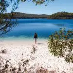 Traveller stands on pristine sandy beach by crystal-clear blue lake during Fraser Experience One Day Tour, enjoying serene lakeside views.