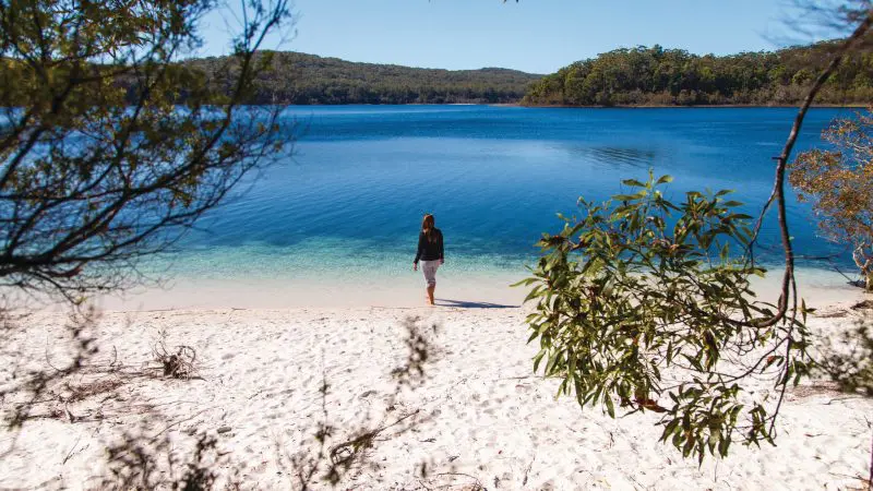 Traveller stands on pristine sandy beach by crystal-clear blue lake during Fraser Experience One Day Tour, enjoying serene lakeside views.