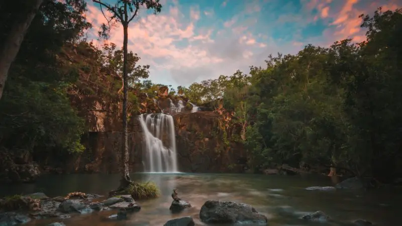 Falls To Paradise Waterfall cascades into a tranquil pool, framed by lush trees and rocks beneath a vibrant sunset, natural beauty.