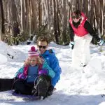 Family sledges down a snowy Lake Mountain hill during their 1 Day Melbourne Snow Waterfalls Tour, smiling in a winter wonderland.