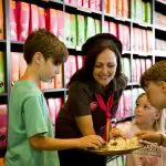 A cheerful woman offers tasty samples to three excited children in a vibrant shop, capturing the excitement of a Lake Mountain Snow Tour.
